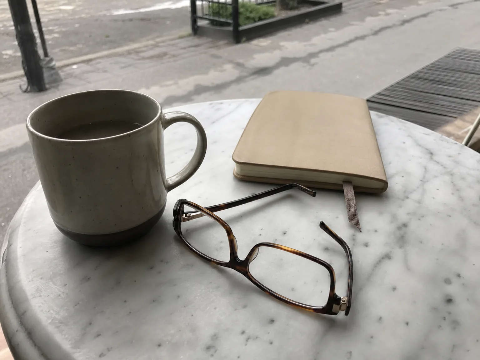 Uma mesa de cafe mostra uma caneca, oculos de leitura e um caderno fechado.