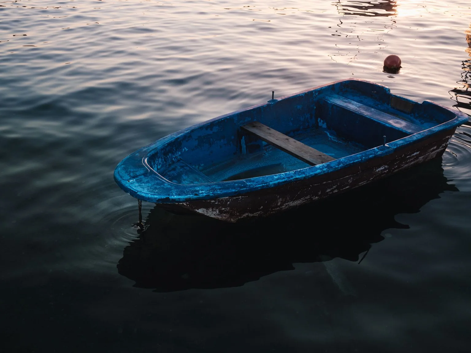 Um pequeno barco azul flutua sobre agua ondulada com luz baixa de fim de tarde.