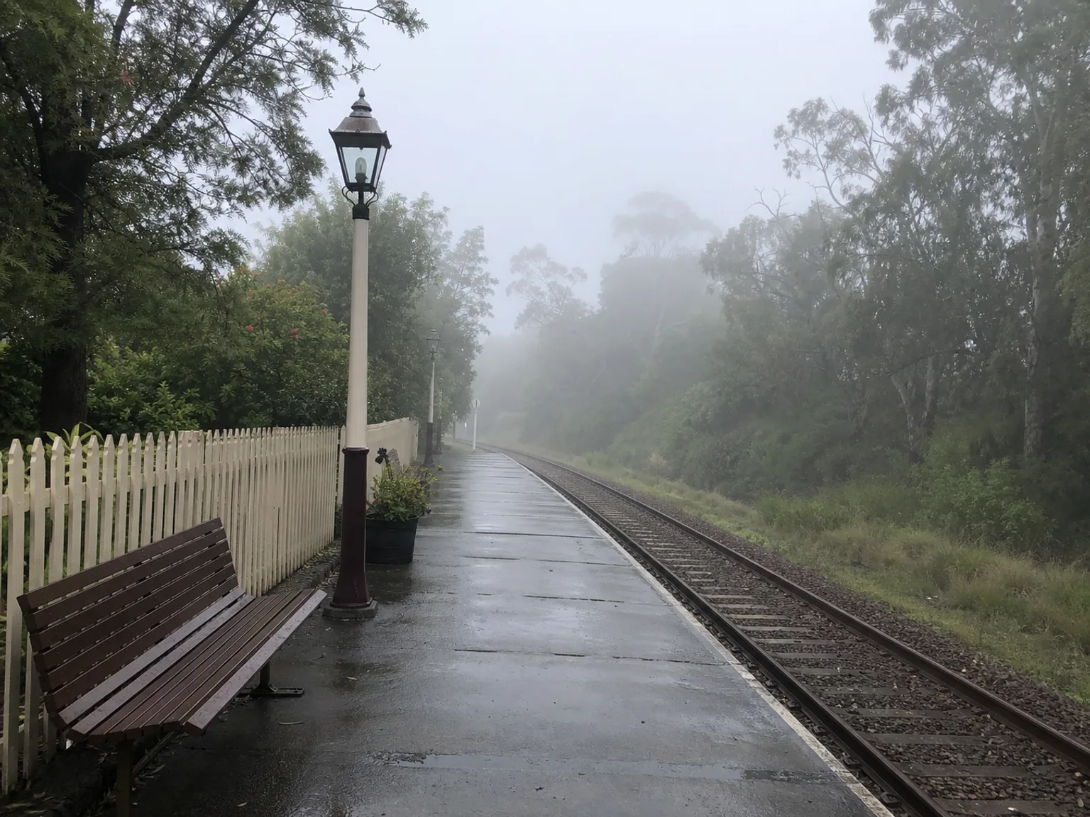 Uma plataforma ferroviaria com nevoeiro aparece junto a carris, bancos e uma vedacao branca.