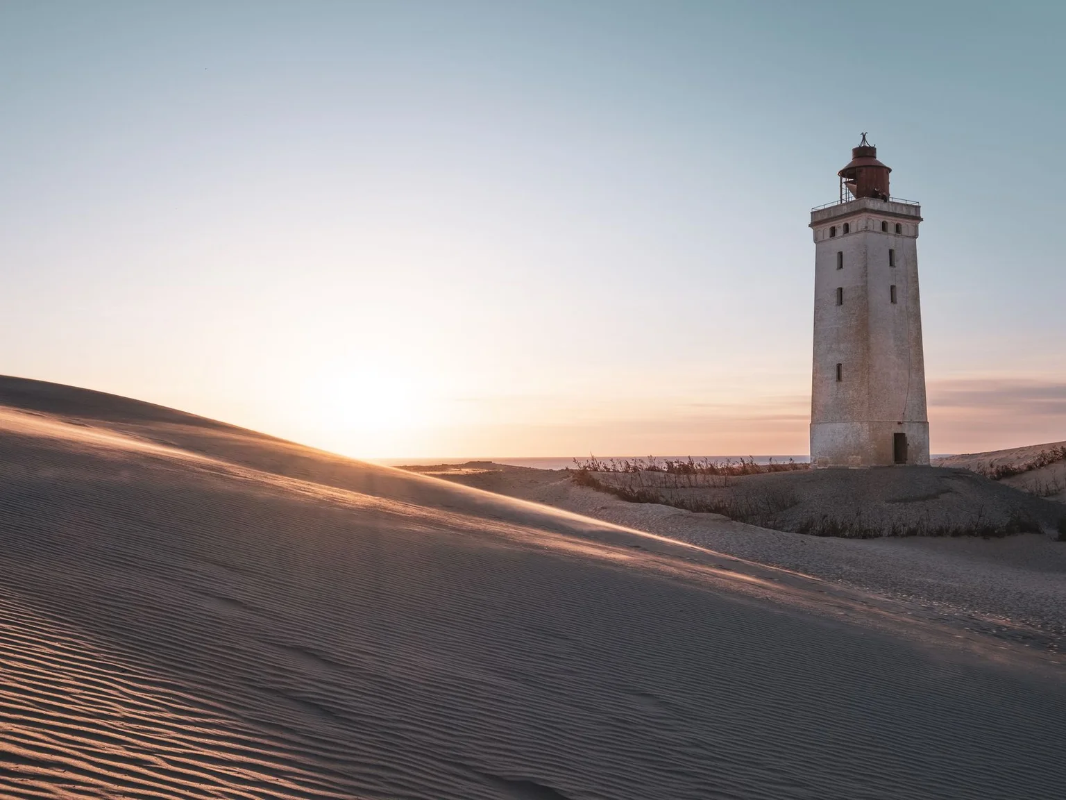 Um farol ergue-se entre dunas enquanto o sol se poe perto do horizonte.