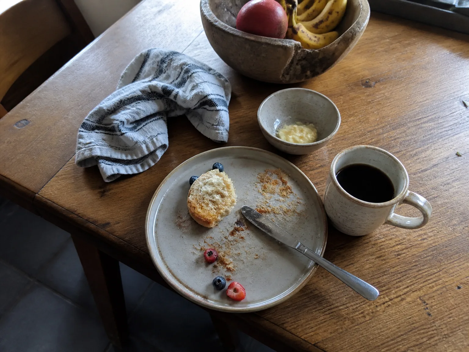 Uma mesa de madeira tem cafe, fruta e restos de pequeno-almoco.