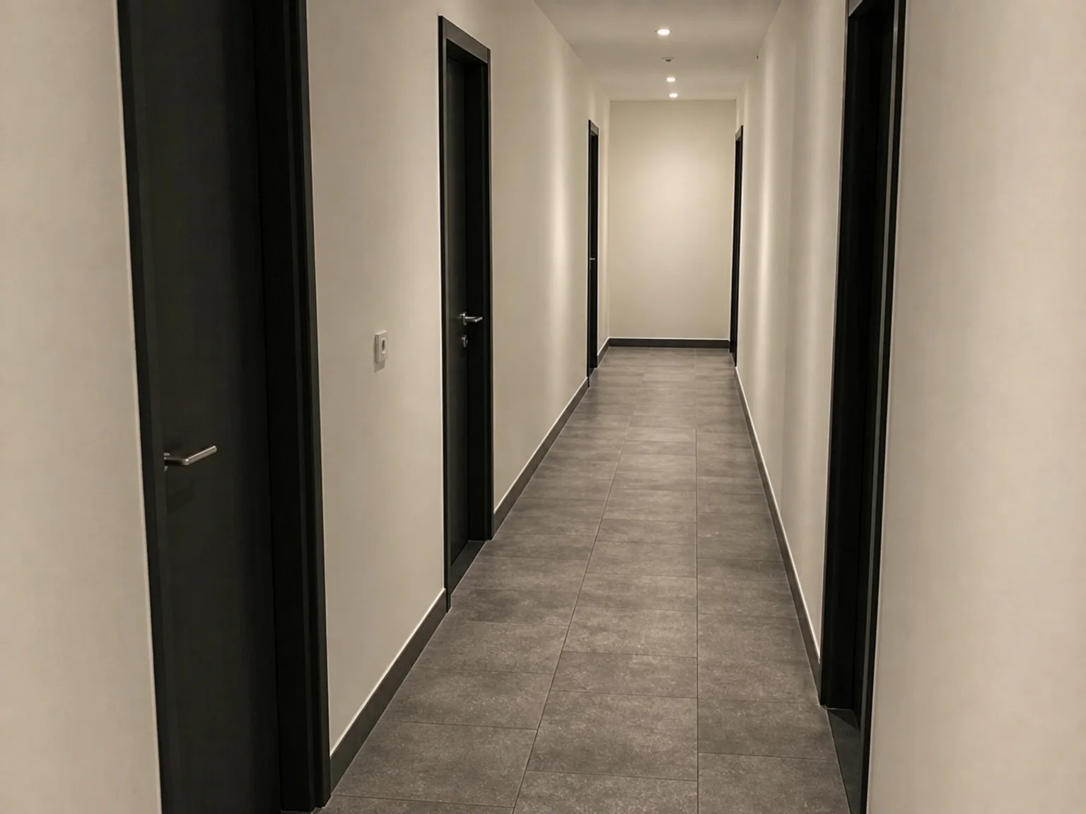 Um corredor longo e vazio esta ladeado por portas escuras e luzes de teto.
