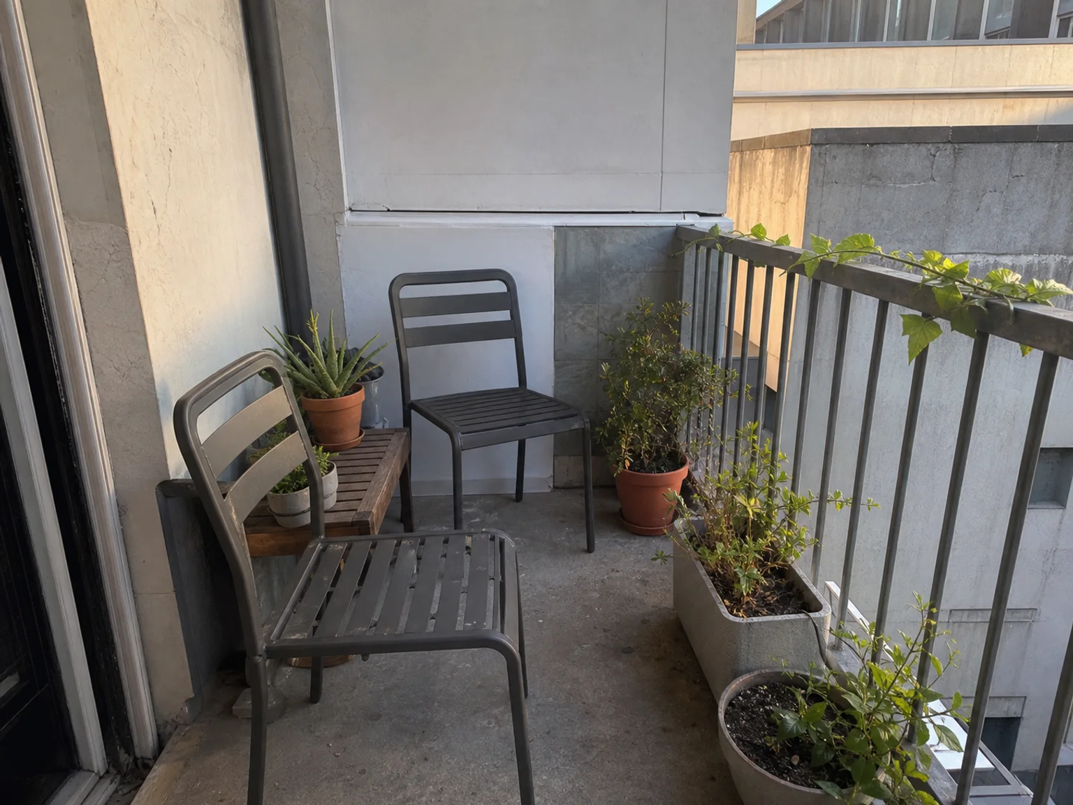 Uma varanda de apartamento tem cadeiras e plantas em vasos num espaco estreito.
